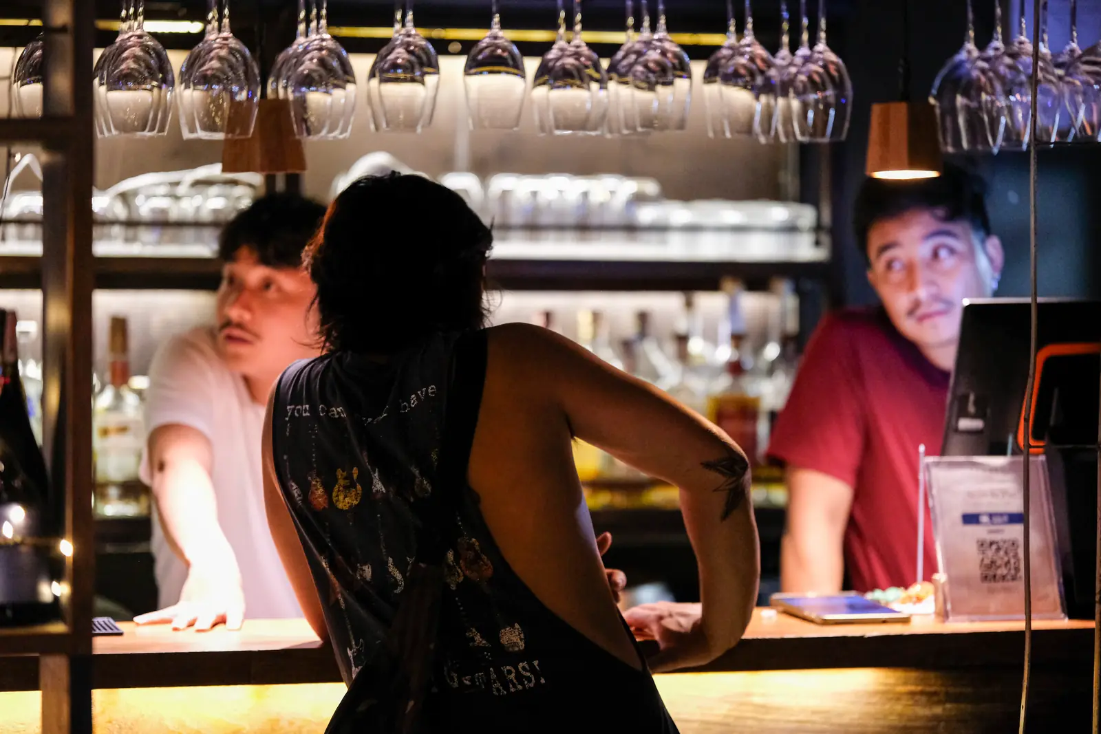 A guest leaning on the counter, talking with the two bartenders under the hanging glasses · Vermut Café, Pai Walking Street