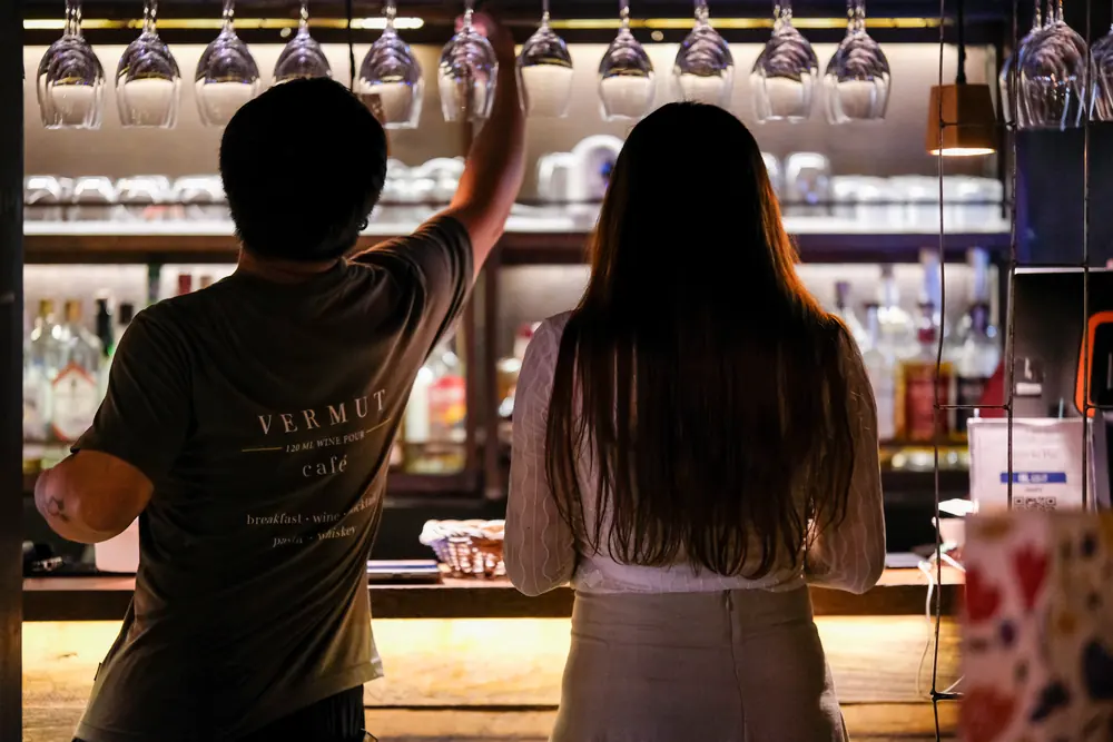 Two of the team reaching for glasses from the overhead rack · Vermut Café, רחוב ההליכה בפאי