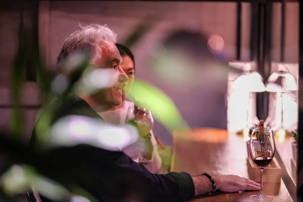 A guest with a glass of red at the front table, seen through the leaves · Vermut Café, Pai Walking Street