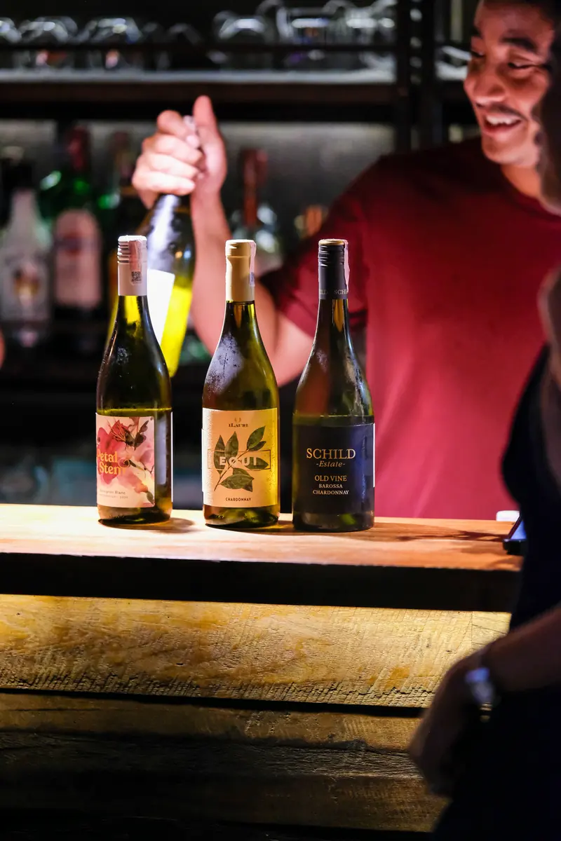 The bartender smiling behind three bottles of white on the counter · Vermut Café, רחוב ההליכה בפאי