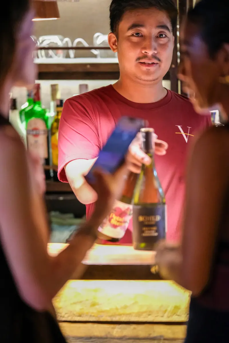 The bartender showing a bottle to two guests at the counter · Vermut Café, רחוב ההליכה בפאי