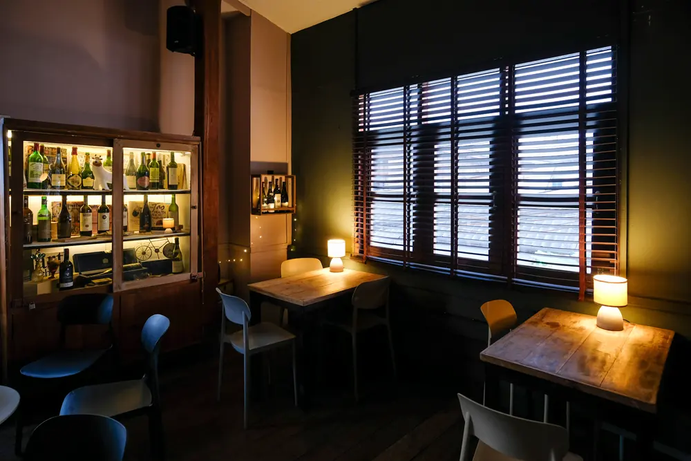 A table by the shutters with a lamp, the wine cabinet beside it · Vermut Café, רחוב ההליכה בפאי