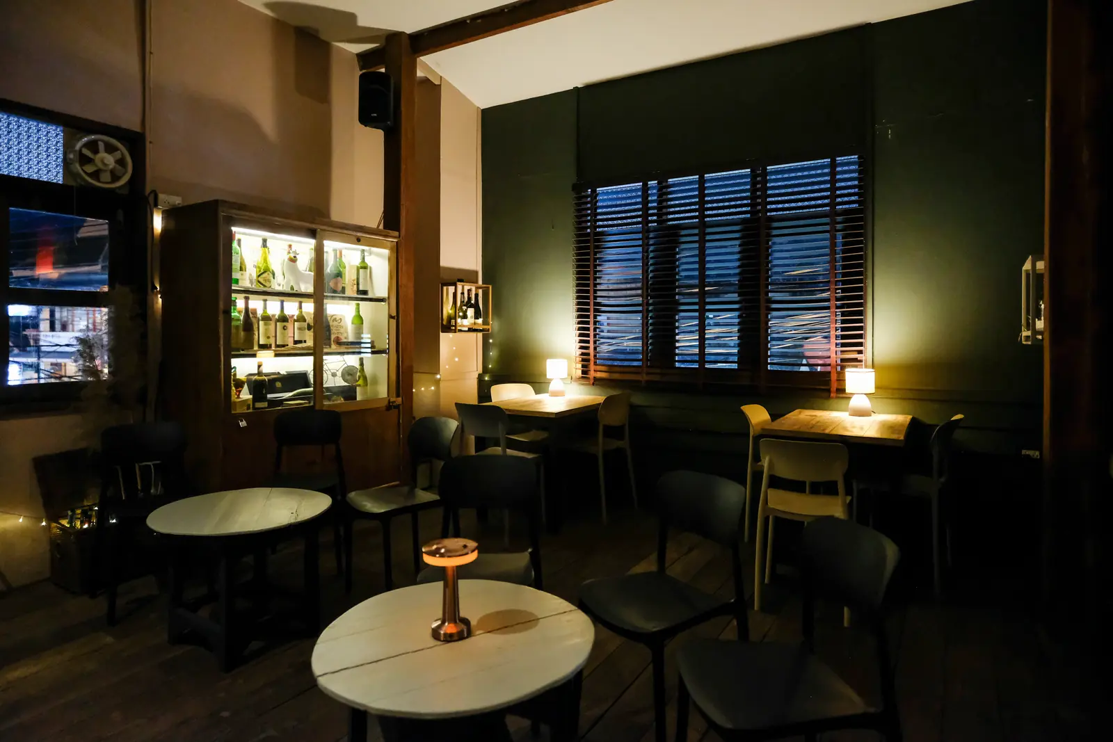 Round tables with lamps under the wooden shutters, the lit wine cabinet in the corner · Vermut Café, Pai Walking Street