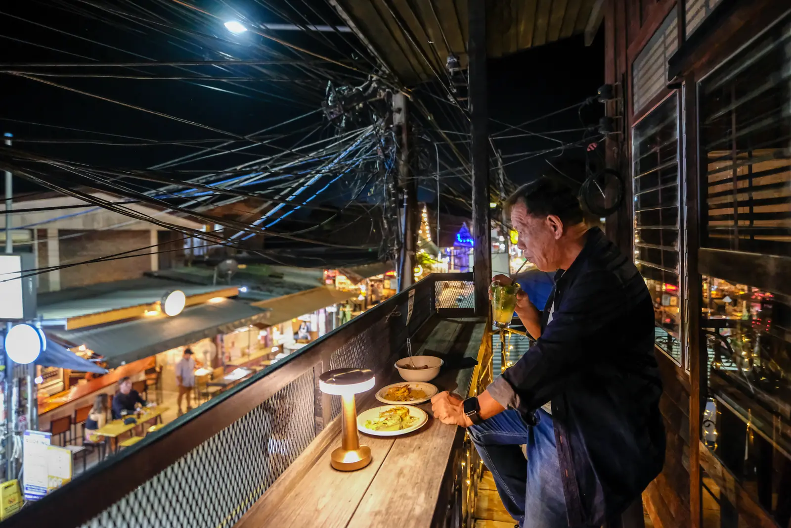 A guest at the balcony rail at night, a plate and a drink beside him, the walking street below · Vermut Café, Pai Walking Street
