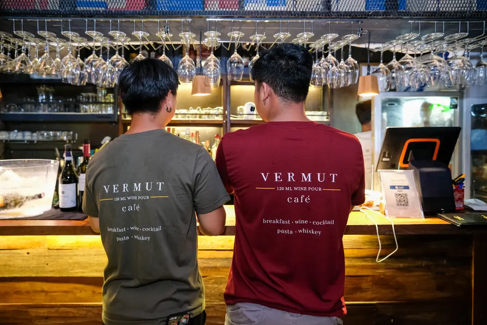 Two of the team at the counter in Vermut café shirts, glasses above · Vermut Café, רחוב ההליכה בפאי