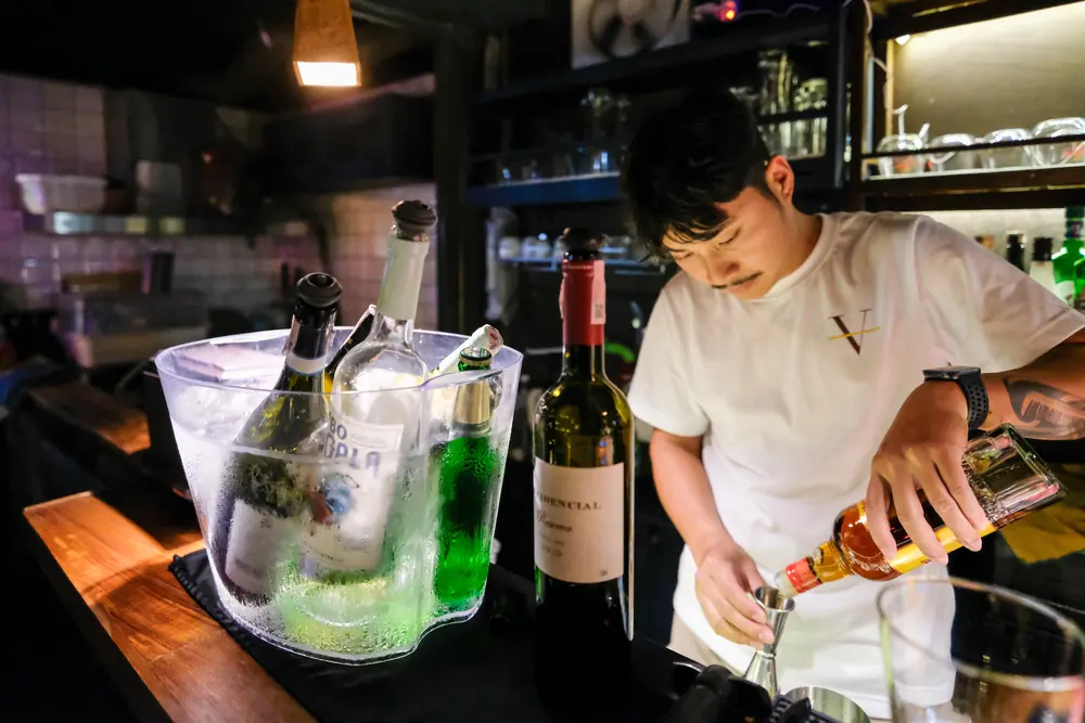 Measuring the pour at the counter, bottles on ice · Vermut Café, רחוב ההליכה בפאי